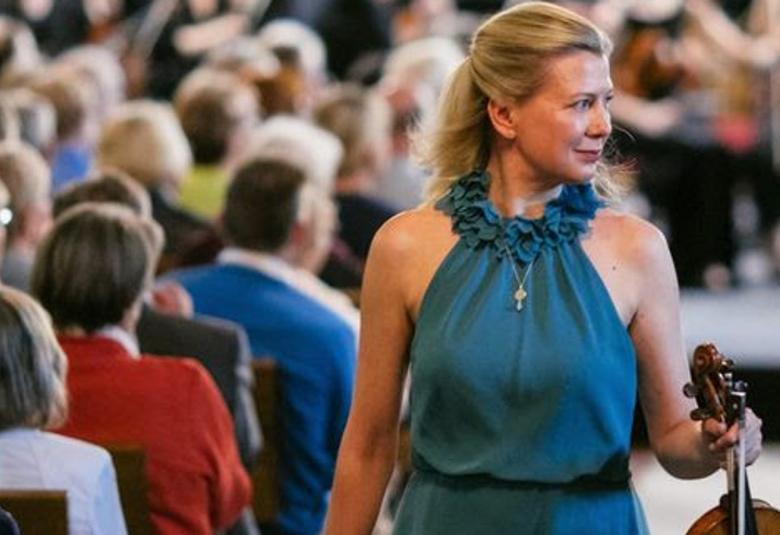 Kuvassa on Elina Vähälä kävelemässä viulu kädessään esiintymästä turkoosissa esiintymisasussa.  Molemmin puolin näkyy yleisöä istumassa.