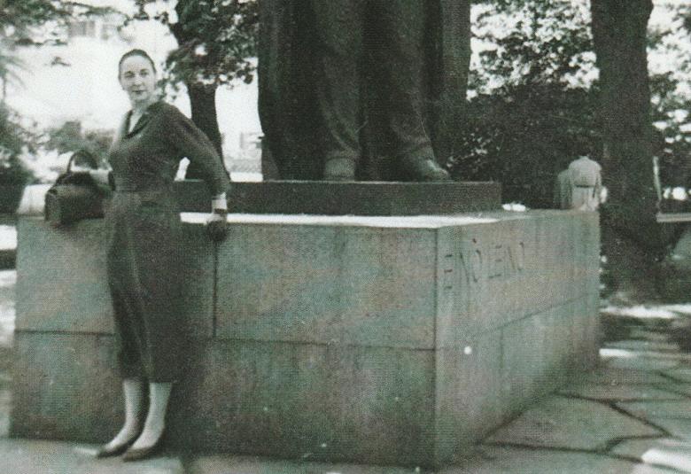 Kuvassa on Helena Eeva ison patsaan juurella seisomassa.  Hänllä on kapea musta puolihame päällään. Kuva on musta-valkoinen.