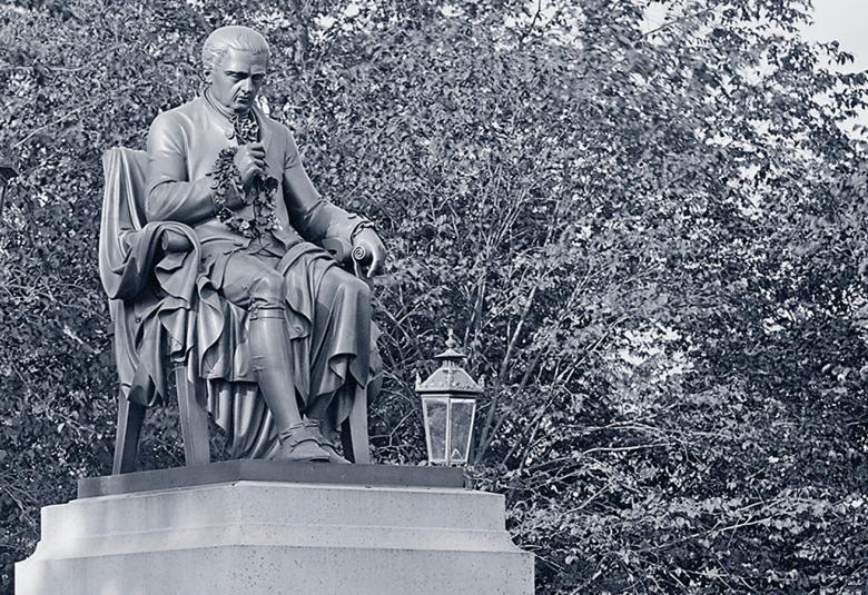 Kuvassa on osa kirjan kannesta, jossa on korkealla jalustalla tuolissa istuvan Porthanin patsas. Kuva on musta-valkoinen.