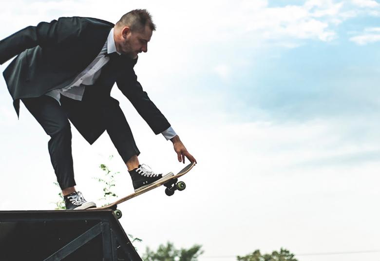 Kuvassa on mies joka on kumartuneena katon harjalla ja pitää vasemmalla kädellään skeittilaudasta kiinni. Taustalla on sini-valkoista taivasta.
