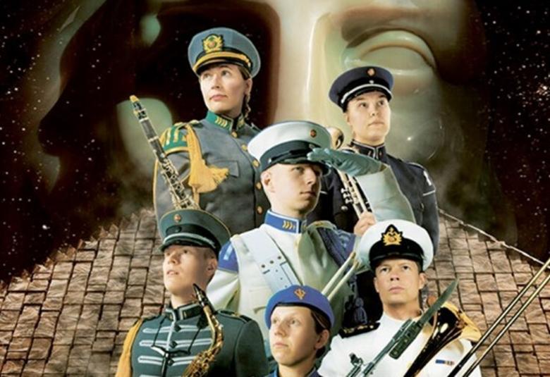 Kuvassa on seitsemän sotilassoittajaa kolmen rivin ryhmässä ja heillä on harmaita tai valkoisia takkeja sekä lakit sekä puhallinsoittimia kädessään. Taustalla on alhaalla tiilikattoa ja takana vaalean patsaan kasvot.   