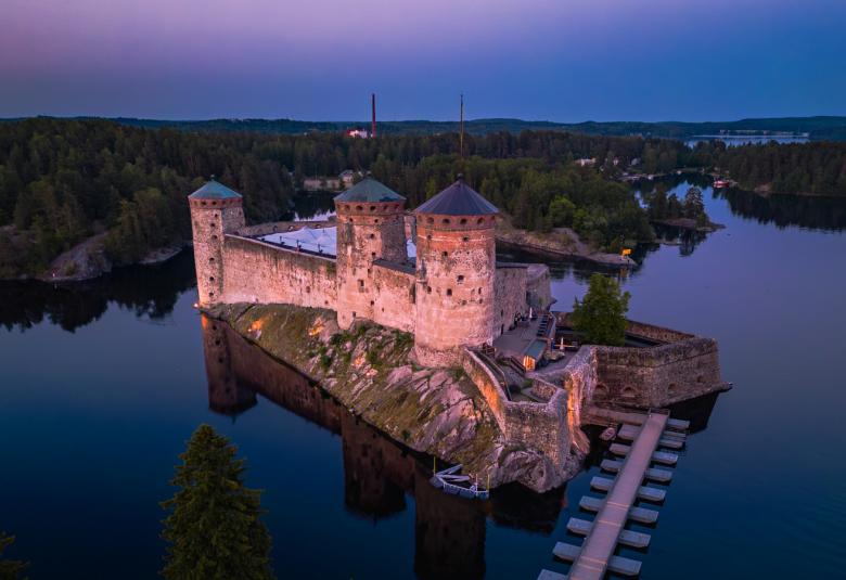 Kuvassa on Olavinlinna sinisävyinen ilmakuva iltahämäräisestä Savonlinnasta, jossa linna on veden keskellä ja taustalla näkyy metsää ja kaupunkia. Edessä on laituria, jossa on paikkoja veneille.