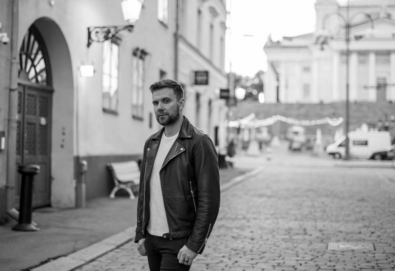 Kuvassa on vanhoja rakennuksia ja katunäkymä katukiveystä.  Antti Ketonen seisoo hieman vasemmalla ja kadulla. Taustalla on Helsingin tuomiokirkko.