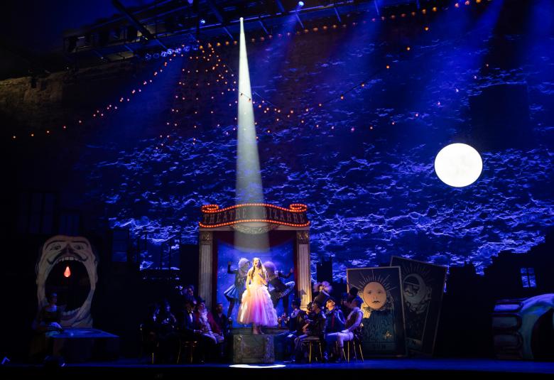 Kuvassa on tummalla ja violetilla valaistulla Olavinlinnan näyttämöllä Lisette Oropesa. Häneen osuu valokeila ja hänellä on vaalea röyhelöinen puku.