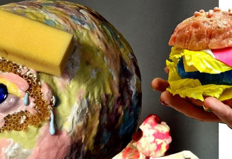 Kuvassa on keltaruskea pää öljyvärillä ja tumman siniset nappisilmät. Kulmakarvat on keltaisina paloina.  Nenässä ja poskissa on vaalean punaista. Oikealla on käsi joka ojentaa värikästä hampurilaista 