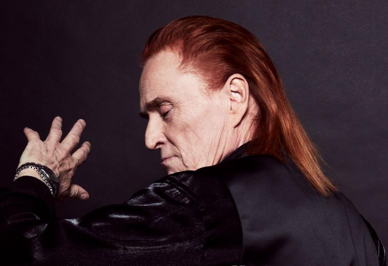 Kuvassa on Tommi Läntinen, joka on selin ja kasvot vinottain vasen käsi kasvojen tasolla. Tausta on musta ja hänellä on musta nahkatakki.