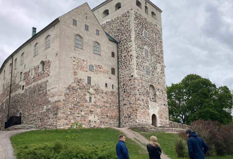 Kuvassa on harmaa Turun linna kuvattuna yläviistoon ja vihreää nurmikenttää.  Alhaalla näkyy kaksi miestä ja nainen katsomassa linnaa.