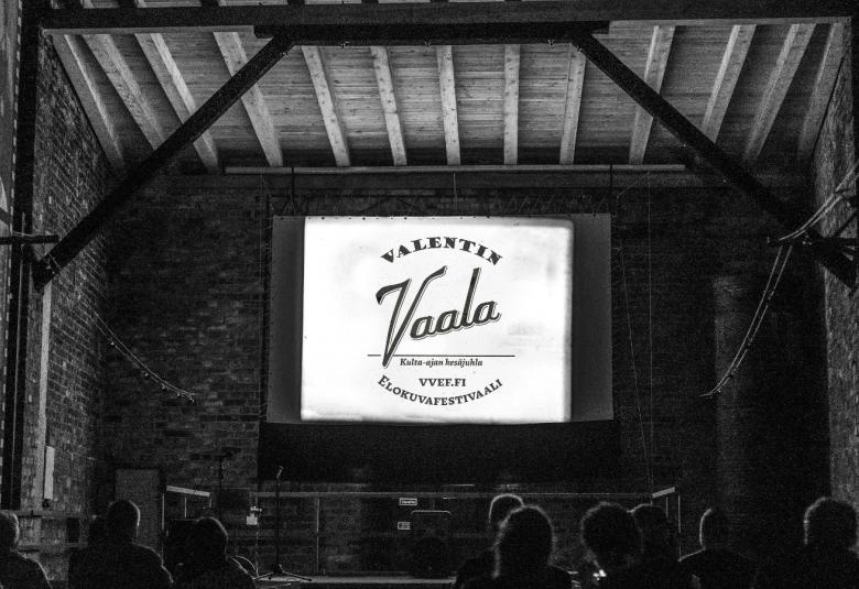 Kuvassa on vanhan ladon katsomo ja lautanäyttämö ja sen keskellä on pieni juliste, jossa lukee Valentin Vaala elokuvafestivaali. Lato on harmaa.