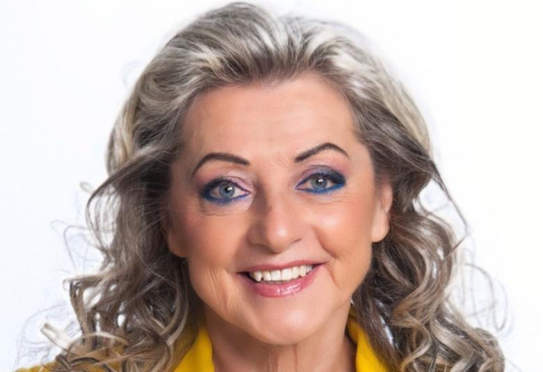 Kuvassa on Kike Elomaa ja hänestä on kasvokuva ja näkyy hieman keltaisen takin kauluksia. Hänellä on vaaleat kiharat hiukset.