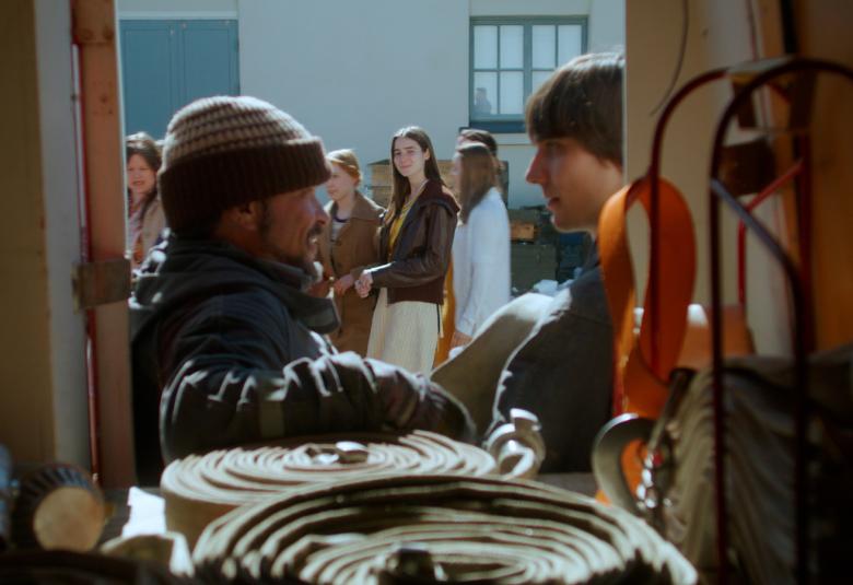 Kuvassa näkyy edessä paloauton sisäosan kautta kaksi palomiestä ja taustalla näkyy ihmisiä ja keskellä nuori nainen.