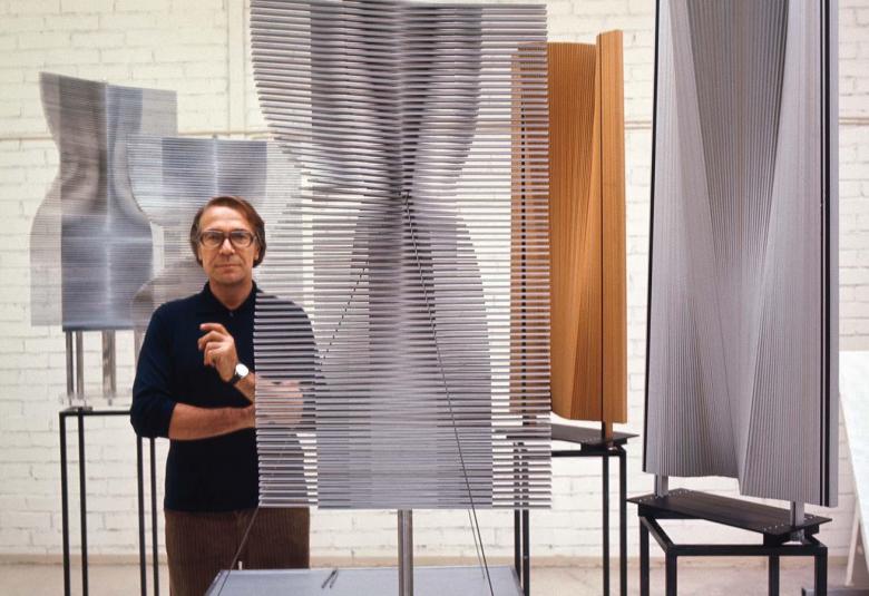 Kuvassa on Raimo Utriainen seisomassa vierellään leijamaisia, kapean metalliraitaisia veistoksia, jotka ovat harmaita.