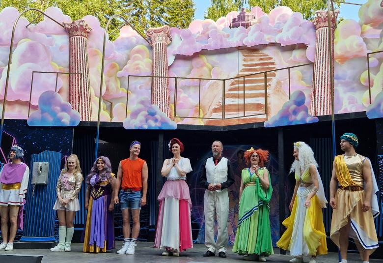 Kuvassa ovat Xanadun näyttelijät lavalla kiittämässä yleisöä. Taustalla on vaalean puna-sinisiä sokeritoppapilviä sekä antiikkisiä pylväitä.