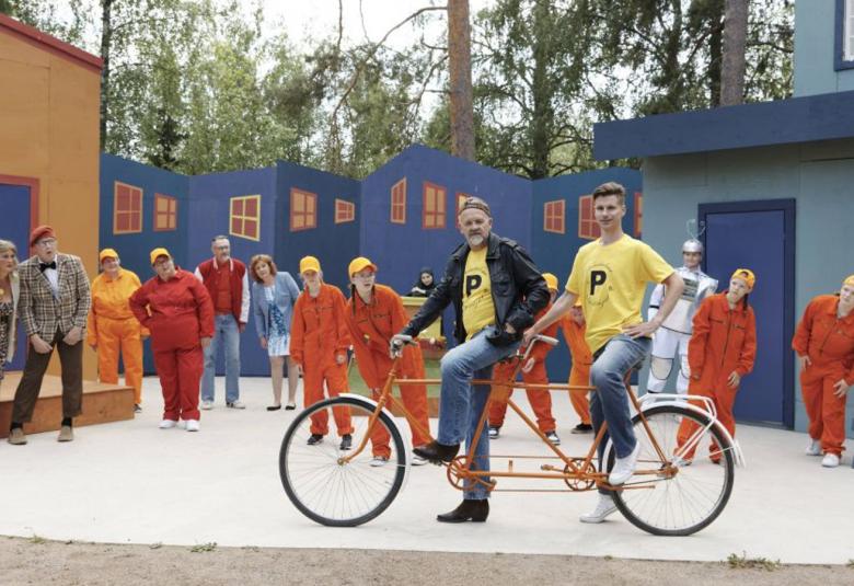 Kuvassa on kaksi miestä tandempyörällä etualalla ja heidän takanaa on pitkä rivi näyttelijöitä katsomassa pyöräilijöitä.