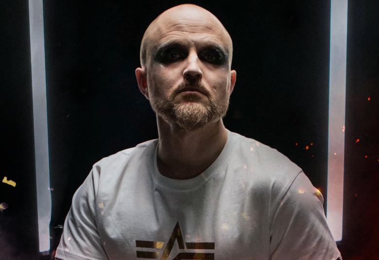 Kuvassa on Esa Arokki istumassa ja hänellä on vaalea t-paita, jossa on keskellä musta kuvio.  Tausta on musta.