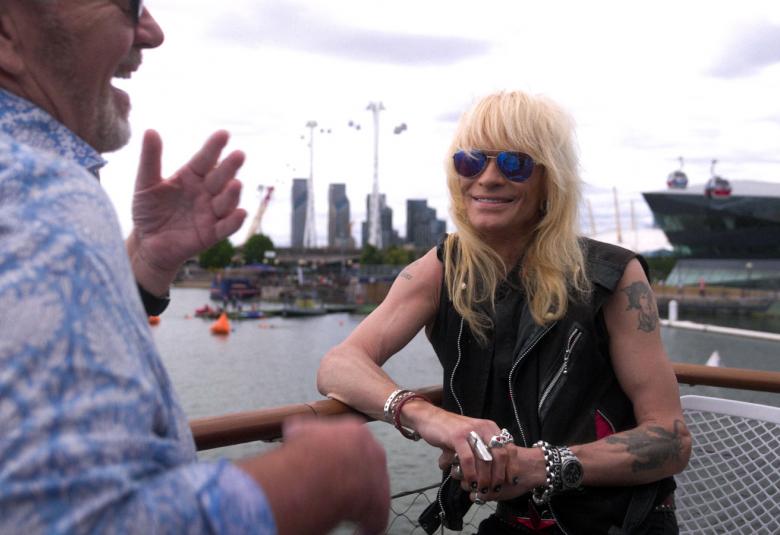 Kuvassa on Michael Monroe istumassa pienessä laivassa kaiteen vieressä. Hän keskustelee selin olevan miehen kanssa. Taustalla näkyy kaupungin silhuettia.