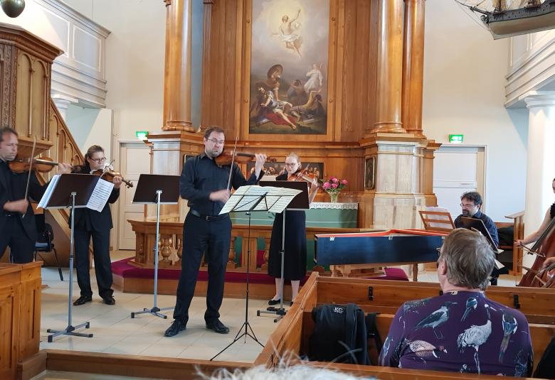 Kuvassa ovat Kaakon Kamarimusiikin ensemblen soittajat Haminan kirkon kuorissa koko kuvan leveydeltä. Taustalla näkyy alttaritaulua ja etualalla kirkon penkkejä sekä oikealla miehen selkämystä.