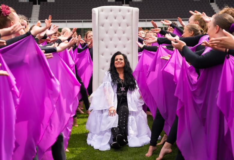 Kuvassa on Kaija Koo keskellä voimistelijoiden kujaa.  Hänellä on valkoinen röyhelöpuku päällään. Voimistelijoilla on päällään mustat trikoot ja ojennetuista käsistä roikkuu violetit huivit.