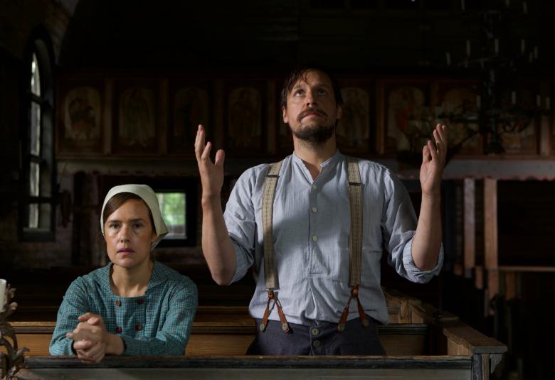 Kuvassa ovat Jessica Grabowsky, joka on pöydän ääressä rukousasennossa polvistuneena ja Jakob Öhman joka on oikealla puolella kädet kohotettuina. Taustalla on hirsitalon pirttiä.