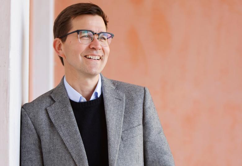 Kuvassa on Paavali Jumppanen puolivartalokuvassa ja hän nojaa valkoiseen seinään.  Jumppasella on vaalea pikkutakki päällään. Taustalla on oranssia seinää.