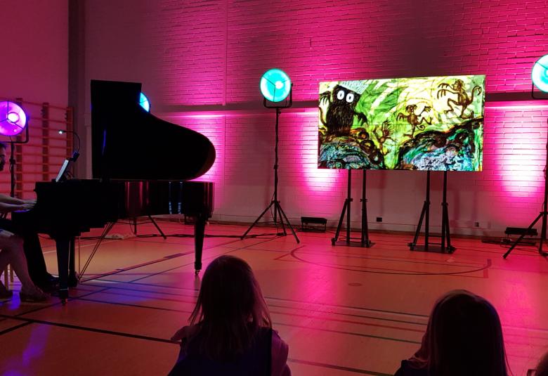 Kuvassa on tummassa salissa vasemmalla flyygeli ja sitä soittamassa Liisa Malmivaara ja Tuomas Turriago. Keskellä värivalojen keskellä on näyttö, jossa on punertavalla pohjalla iso kuningashahmo ja vieressä peikkoja.