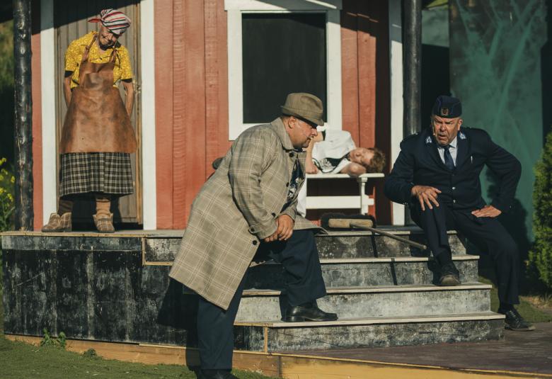 Kuvassa on punainen mökki, jonka portailla istuu oikealla Reinikainen ja vasemmalla mies, jonka vasen jalka on portaalla ja hän nojaa siihen keskustellessa Reinikaisen kanssa. Vasemmalla mökin kulmalla on nainen ruskeassa mekossa.