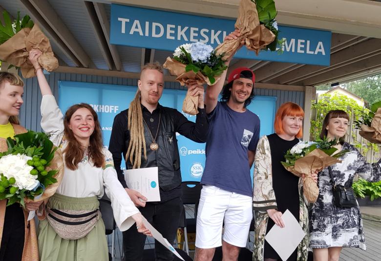 Kuvassa ovat palkitut taiteilijat ja he tuulettavat kukkien kanssa iloisesti. Vasemmalta Juliana Hurri, Kateryna Yehrova, Sakari Vinko, Sergii Shaulis, Melissa Sammalvaara ja Oksana Mykhanko. Taustalla on kyltti vaalean sinisellä pohjalla valkoisin kirjaimin Taidekeskus Salmela.