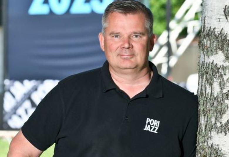 Kuvassa on Sampsa Jolma nojaamassa koivun runkoon ja hänellä on tumman sininen Pori Jazz -t-paita päällään.  Taustalla näkyy iso Pori Jazz 2023 kyltti.