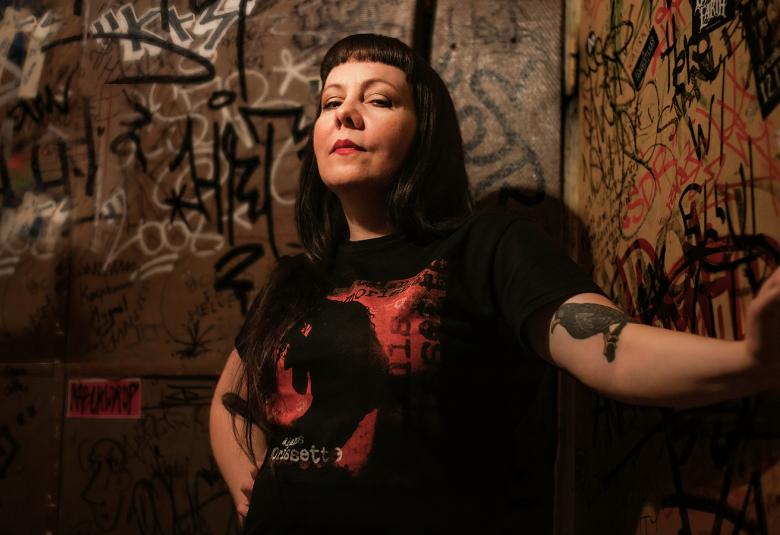 Kuvassa on Tiina Käpylä ja hänellä on tummat, pitkät hiukset sekä musta-punainen puku. Taustalla on graffitiseinää.