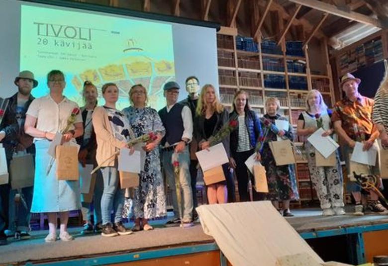 Kuvassa ovat lavalla kirjoituskilpailun palkitut 20 osallistujaa. Heillä on palkintokirjat kädessään. Taustala on heijastettu kirjoituskilpailun nimi Tivoli.