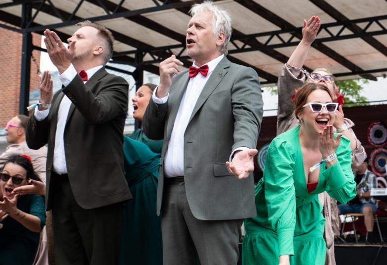 Kuvassa on lavalla lalajia ja edessä kaksi miestä mikkien kanssa laulamassa sekä oikealla vihreässä puvussa nuori nainen.