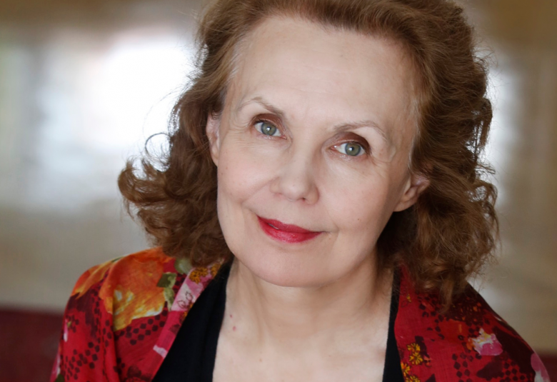 Kuvassa on Kaija Saariaho puolivartalokuvassa ja hänellä on punakirjavassa jakussa. Kuva on otettu hieman yläviistosta.