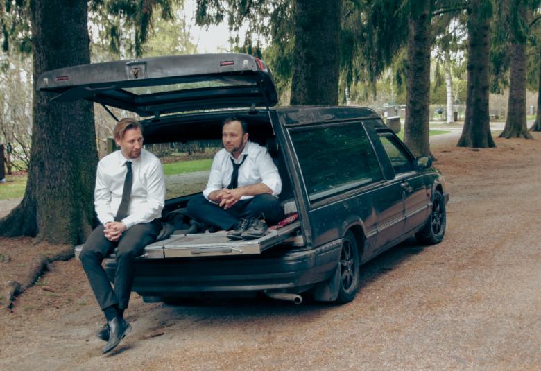 Kuvassa ovat Pekka Strang ja Jari Virman istumassa ruumisauton avonaisella takaluukulla. Auto on parkeerattu mäntykujalle hautausmaalla.