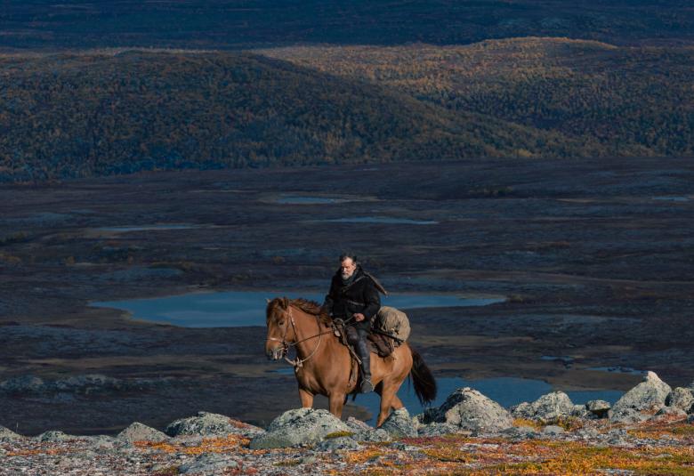 Kuvassa on ratsastaja tunturimaisemassa ja tausta on tumman sininen.  Sieltä näkyy tunturin ääriviivaa.  Ratsastaja kulkee kivistä tunturin lakea.