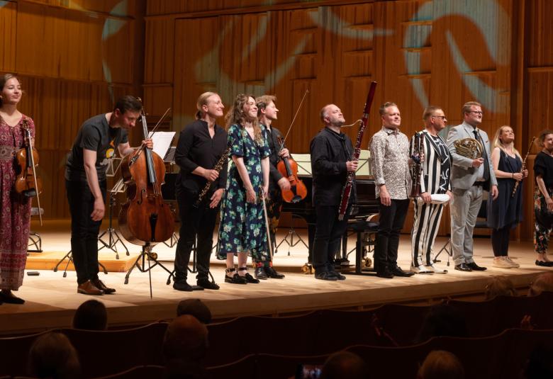 Kuvassa soittajat seisovat esiintymislavalla ja kiittävät.  Heitä on lukuisia. Valaistus on himmeä.