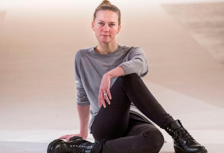 Kuvassa on Auri Ahola istumassa lattialla toinen polvi nostettuna ylös ja vasen käsi polven päällä. Hänellä on harmaa pusero ja mustat housut päällään sekä mustat kengät.