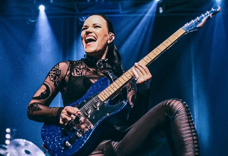 Kuvassa on Erja Lyytinen soittamassa kitaraa.  Hänellä on vasen jalka tuolilla ja hänellä on mustat vaatteet päällään.  Tausta on sininen ja ylhäältä päin tulee kaksi valoa.