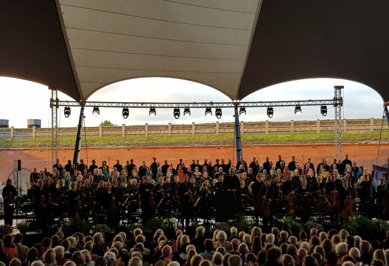 Kuvassa ovat kuorot ja orkesteri sekä edessä yleisöä. Taustalla on Bastionin tiilimuuria ja nurmea. Päällä on pingotettua musta-valkoista telttakattoa.