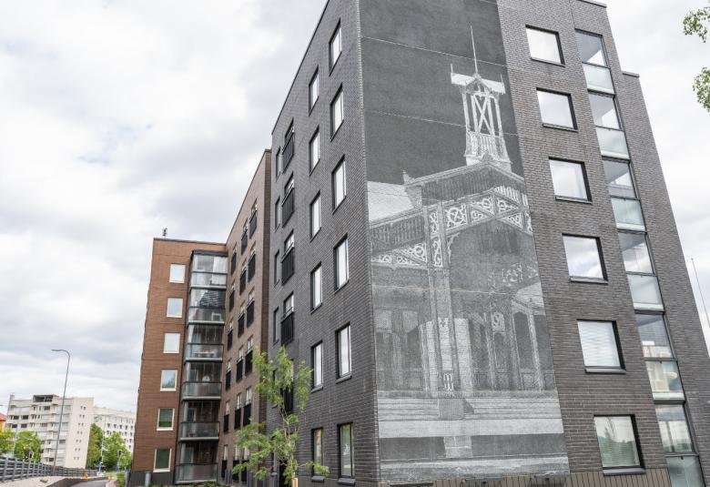 Kuvassa on iso harmaan kerrostalon päädyssä harmaalla vanhan Lahden rautatieaseman piirros. 