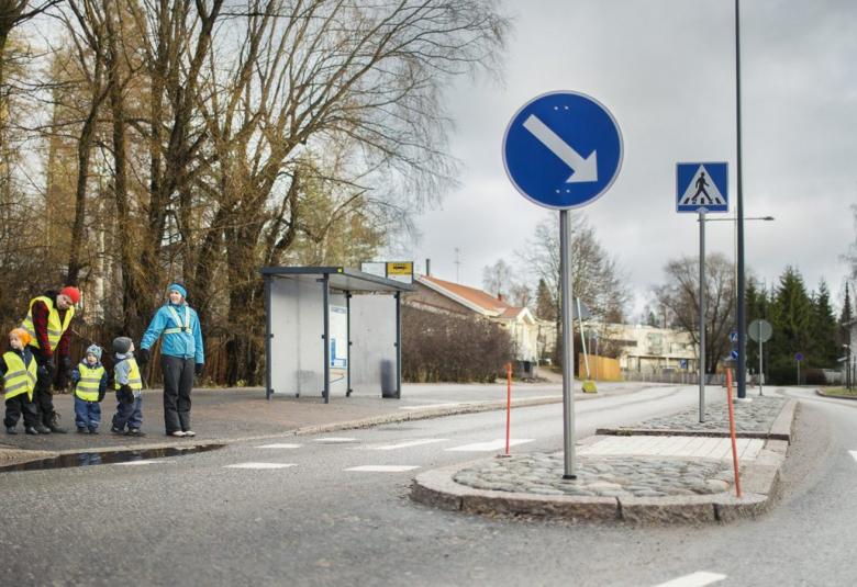 Kuvassa on vasemmalla lähdössä ylittämään katua keltaisessa liivissä nainen, jolla on kaksi pientä keltaliivistä lasta kädessään. Vieressä nainen vaalean sinisessä heidastinnauhaisessa takissa ja pitää kädestä pientä lasta. Keskellä kuvaa on liiktenteen jakaja.