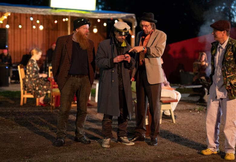 Kuvassa neljä miestä etualalla katsomassa yhden kännykkää.  Taustalla on talo ja edessä istuu ihmisiä tuoleilla.  Kuva on otettu illalla ja on pimeää.