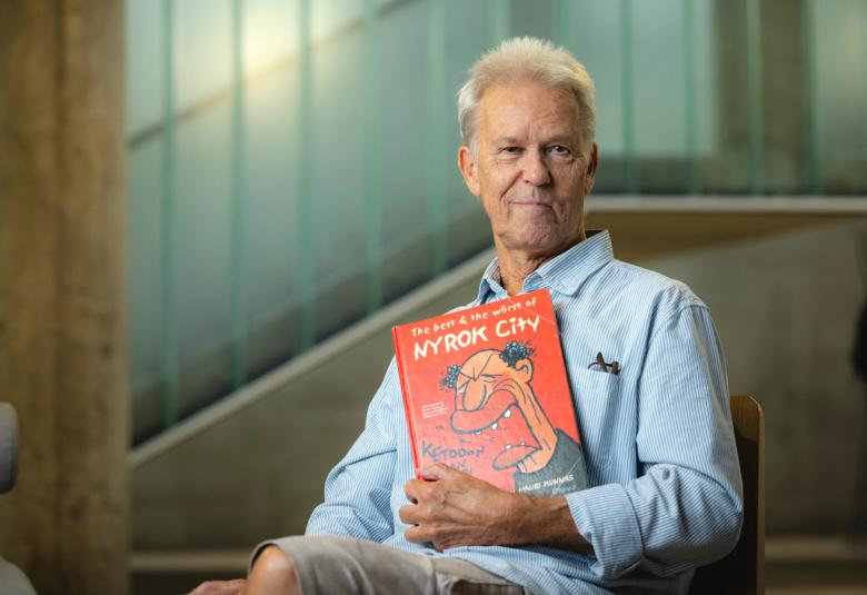 Kuvassa on Mauri Kunnas istumassa ja hän näyttää kirjaansa. Hänellä on vaalean sininen paita päällään.  Taustalla on pitkulaisesti ruudutettua sinertävää lasia ruskeilla puisilla tukipuilla.