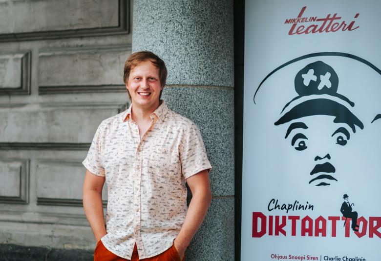 Kuvassa on Juha Pulli, joka nojaa isotiiliseen Mikkelin teatterin seinään.  Oikealla puolella on Diktaattorin juliste, joka on musta-valkoinen ja siinä on hitlermäinen graafinen kasvokuva sekä näytelmän nimi.