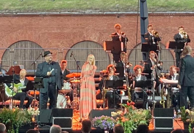Kuvassa on orkesterin jäseniä ja keskellä laulamassa Sami Pitkämö ja Aili Ikonen. Taustalla on bastionin tiiliseinää ja siinä varastojen puisiä ovia.