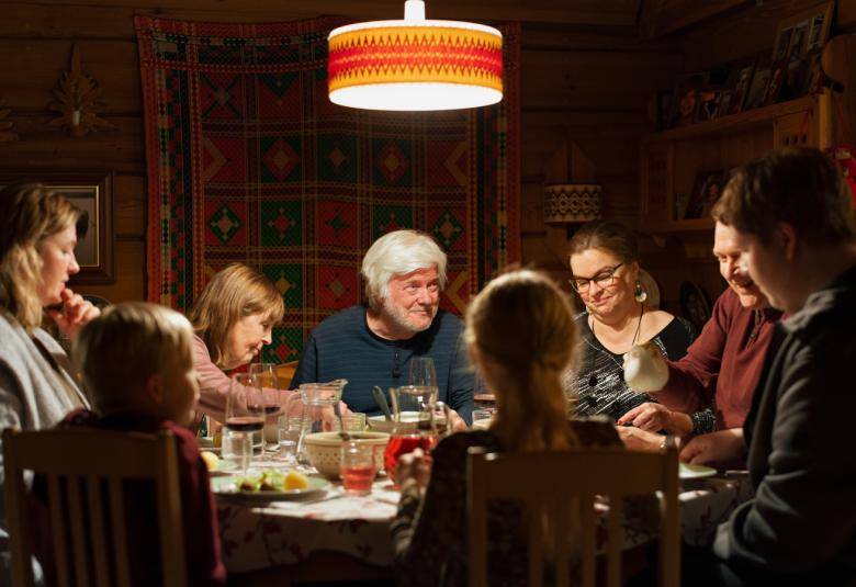 Kuvassa on keskellä pöydän ääressä vaalea hiuksinen vanhempi mies ja ympärillä eri-ikäisiä henkilöitä kuusi ja yksi lapsi syömässä.  Taustalla oleva seinä on ruskea.