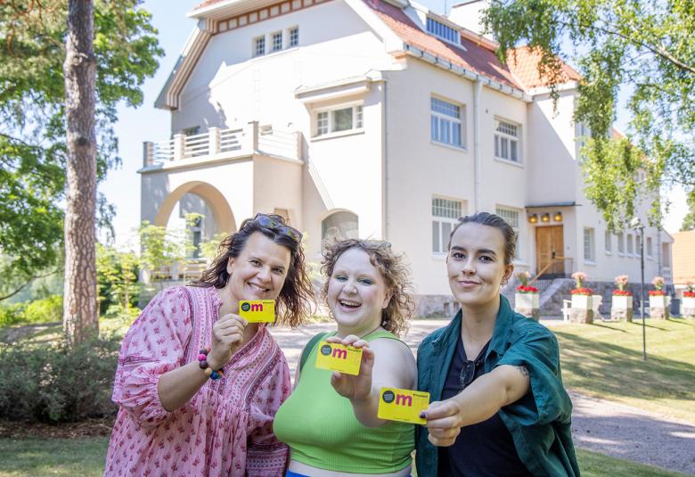 Kuvassa on taustalla Tamminiemen päärakennus, joka on vaalea jugend-talo.  Edustalla on kolme naista vierekkäin ja he näyttävät kaikki museokorttia.