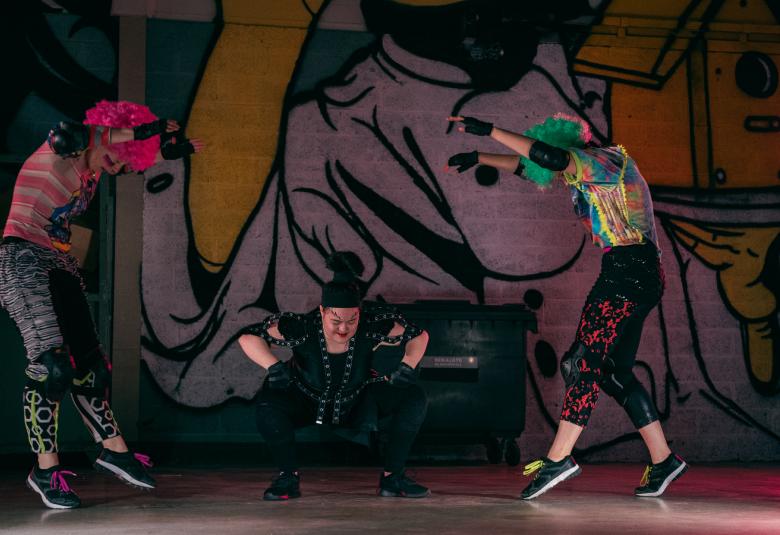 Kuvassa on tummalla kuviollisella taustalla kaksi tanssijaa molemmilla puolilla hyppäämässä selät köyristyneinä ja keskellä istumassa yksi tanssija. 