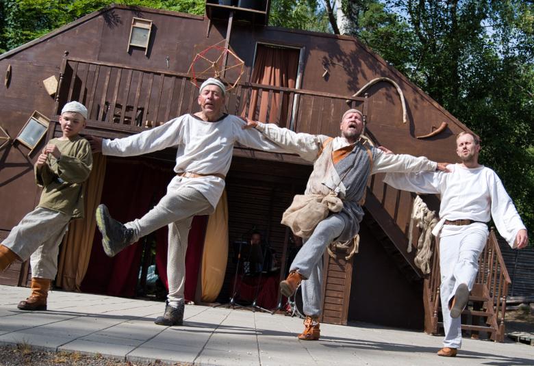 Kuvassa on vasemmalla nuori poika vaaleissa housuissa ja vihertävässä puserossa ja hänestä oikealle on kolme miestä vaaleissa housuissa ja puseroissa.  He kaikki tanssivat ja oikeat jalat on viety vasempaan suuntaan.  Taustalla on tumman ruskea talokulissi.