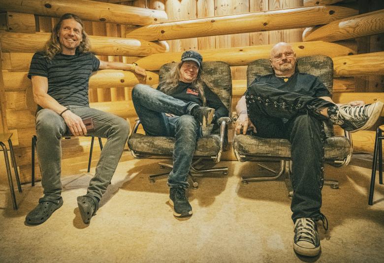 Kuvassa ovat Tipe Johnson, Pete Kiisseli ja Ville Soininen istumassa rennosti tilassa, jossa on poikittain pyöreitä hirsiä ja kapeampia pystyrimoja lautaseinän päällä.  Lattia on vaaleaa mattoa. Miehillä on tummat vaatteet päällään.