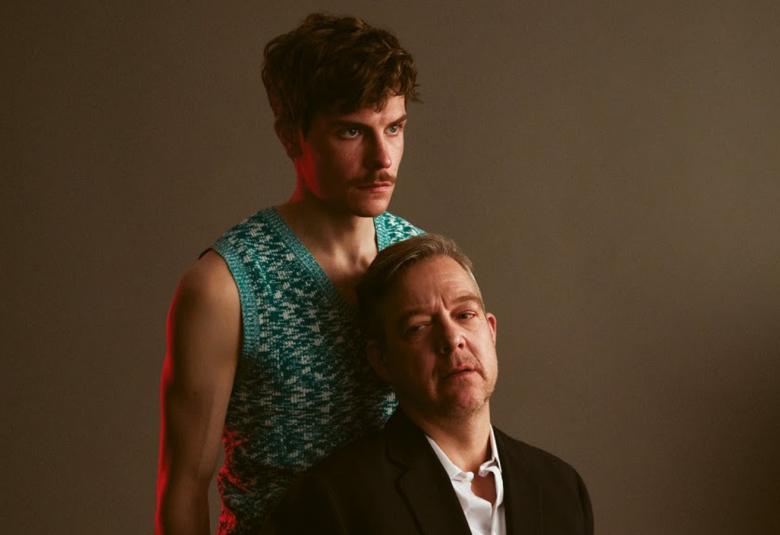 Kuvassa ovat Riko Eklundh seisomassa takana sinertävässä liivissä ja Mikko Kauppila alempana edessä istumassa ja häenllä on musta takki. Tausta on tummahko.