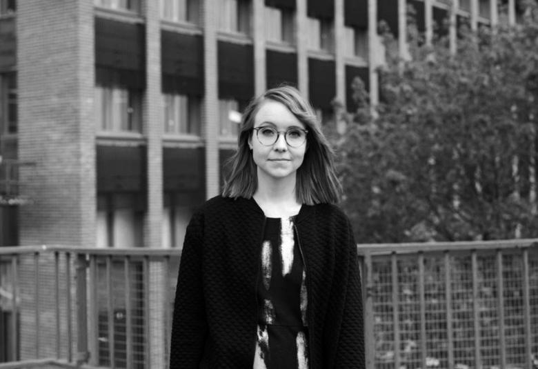 Kuvassa on Riikka Manninen seisomassa taustallaan toimistotalo. Kuva on musta-valkoinen.